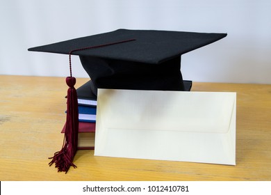 Black Bonnet For Graduation Ceremony On Top Of Books With Envelope On The Right Side.