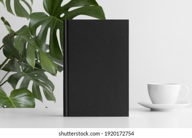 Black Book Mockup With A Cup Of Coffee On A White Table And A Monstera Plant.