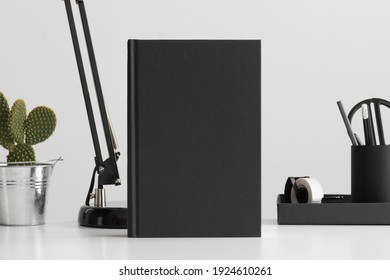 Black Book Mockup With Workspace Accessories And A Cactus On A White Table.