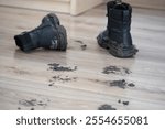 Black boots sit next to muddy footprints on a polished wooden floor. Uncleaned footprints on wooden floor after outdoor activity. Concept of dirty stains and house cleaning.