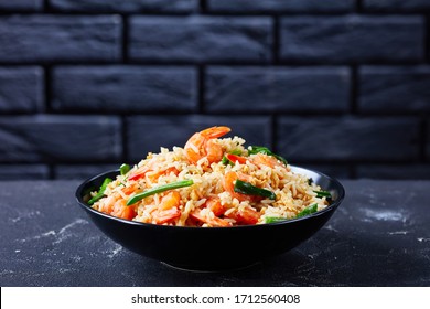 Black bowl with asian fried jasmine rice with shrimps scrambled eggs and spring onions with thai sauce on a dark concrete background in front of black brick wall, close-up, copy space
