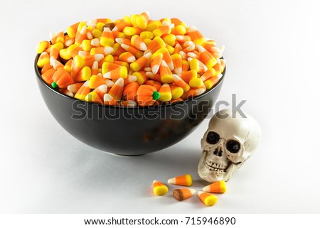 A black bowl over filled with Candy Corn taken on a white background.  A plastic toy skull is also shown in the photo