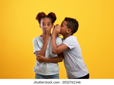 Black Boy Brother Sharing Secrets With His Older Sister, African American Teen Guy Whispering Something To Cute Girl Sibling, Yellow Studio Background. Brotherhood, Sisterhood, Friendly Family Concept