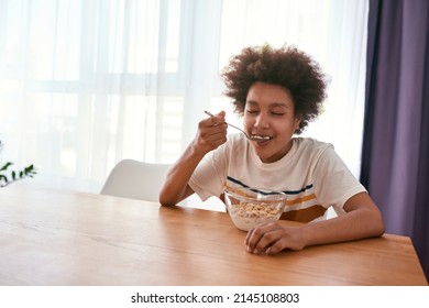 Black Boy With Closed Eyes Enjoying Eating Cereals At Wooden Table At Home. Curly Teenager Child Having Breakfast In Sunny Morning Time. Domestic Lifestyle. Healthy Eating. Spacious Moden Apartment