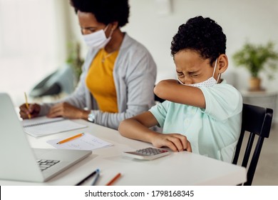 Black boy coughing into elbow while wearing face mask during home schooling due to coronavirus pandemic.  - Powered by Shutterstock