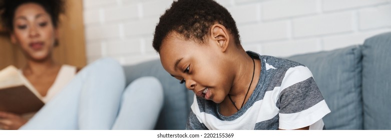 Black Boy Drawing And Sitting On Sofa While His Mother Reading Book At Home