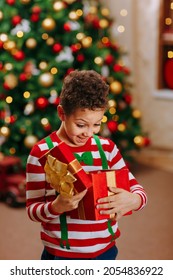 A Black Boy In A Striped Red And White Jacket Holds And Open A Red Box With A Christmas Gift, On The Christmas Tree Background.