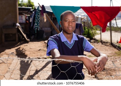 Black Boy With A Very Serious Expression Standing Against The Fence In A Township.