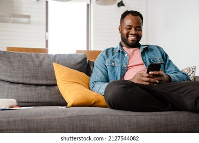 Black Bristle Man Smiling And Using Cellphone While Sitting On Sofa At Home