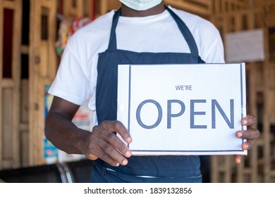 Black Business Owner Wearing A Face Mask And Holding An Open Sign In Front Of His Shop