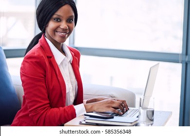 Black Business Woman Looking At Camera While Smiling, With Her Hands Still On The Keyboard Of Her Laptop Computer Where She Was Sitting Working Before Looking Up At Camera.