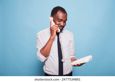 Black businessman holding receiver to ear and talking on landline telephone while listening to person speaking at other end of call. African American employee in white shirt using old school phone. - Powered by Shutterstock