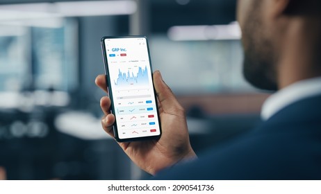 Black Businessman Holding Smartphone and Checking Stock and Cryptocurrency Market in Office. African-American Businessperson using Internet with Mobile Phone Device. Over Shoulder Shot - Powered by Shutterstock