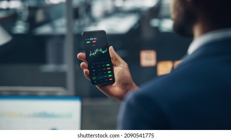 Black Businessman Holding Smartphone And Checking Stock And Cryptocurrency Market In Office. African-American Businessperson Using Internet With Mobile Phone Device. Over Shoulder Shot