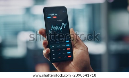 Black Businessman Holding Smartphone and Checking Stock and Cryptocurrency Market in Office. African-American Businessperson using Internet with Mobile Phone Device. Over Shoulder Shot
