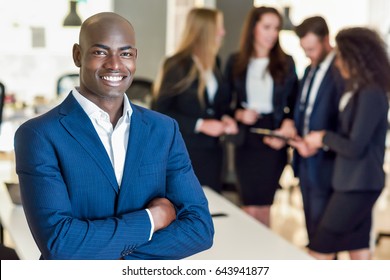 Black Businessman Leader Looking At Camera In Modern Office With Multi-ethnic Businesspeople Working At The Background. Teamwork Concept. African Man Wearing Blue Suit.