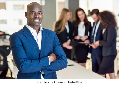 Black Businessman Leader Looking At Camera In Modern Office With Multi-ethnic Businesspeople Working At The Background. Teamwork Concept. African Man Wearing Blue Suit.