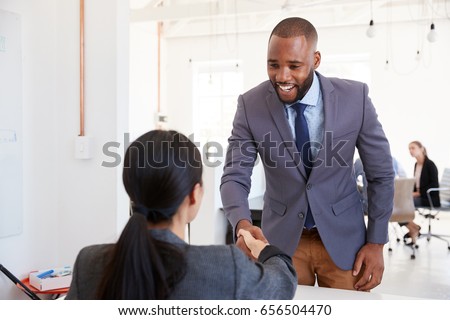 Black businessman and seated woman shaking hands in office