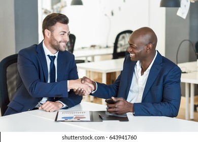 Black Businessman Shaking Hands With A Caucasian One Wearing Suit In A Office. Two Smiling Men Wearing Blue Suits Working In An Office With White Furniture