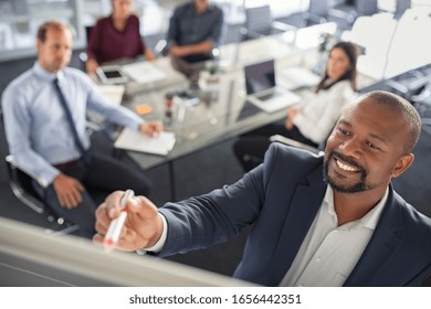 Black Businessman Writing On A Whiteboard While Explain New Strategy. Mature Entrepreneur Discussing Business Ideas And Plans With Colleagues In Background. Top View Of Happy African American Leader I