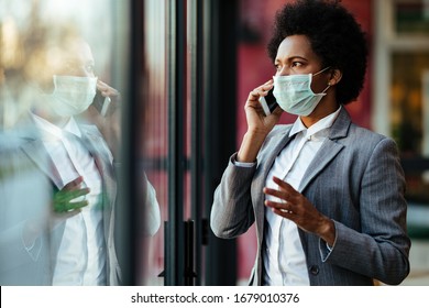 Black businesswoman wearing protective mask on her face and talking on smart phone while looking through the window.  - Powered by Shutterstock