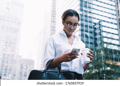 Black busy serious brunette in glasses messaging on mobile phone carrying black bag and holding cup of coffee with modern city on background