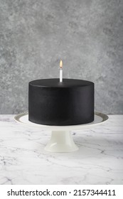 Black Cake With A Candle On A Cake Stand, White Table.