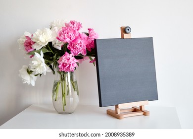 Black Canvas Mockup On Wooden Easel And Vase With Pink Flowers On Table On White Wall Background. Blank Artistic Canvas