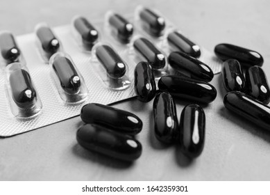Black capsule pills in a blister pack and scattered on a light gray background.