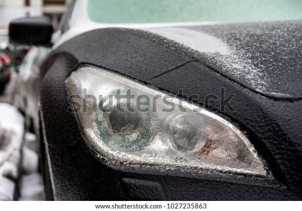 Black car coated ice crust\
during the  icy rain in cold weather. Front part of the car\
headlights