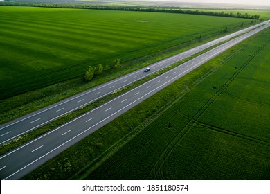 Black Car Driving On Asphalt Road Along The Green Fields. Seen From The Air. Aerial View Landscape. Drone Photography.  Travel Concept. Sunset Time