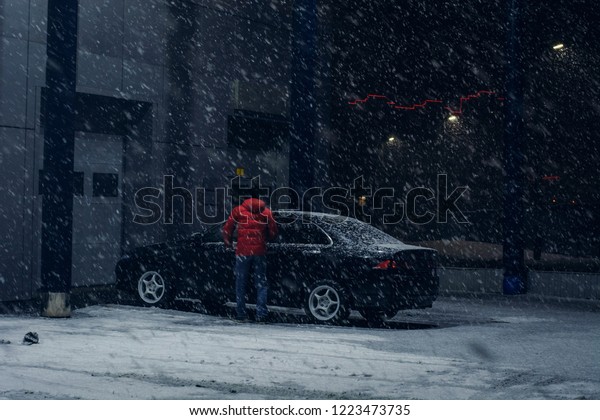 Black car parked\
near office building in winter evening in strong snow storm.\
Automobile near auto service in night. Driver near machine near\
automatic gate for\
maintenance.