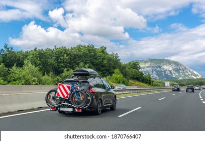 Black Car With Roof Luggage Box And Trunk Bike Rack Driving On The Highway. Beautiful Mountain Landscape Background