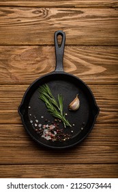 Black Cast Iron Frying Pan Skillet With Food Cooking Ingredients Fresh Rosemary, Garlic, Salt And Pepper On Rustic Wooden Table, Cooking Background And Healthy Eating Kitchen Concept 