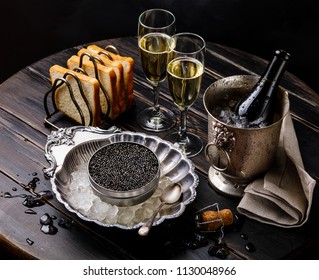 Black caviar in can on ice in silver bowl, bread and champagne in ice bucket on black wooden background