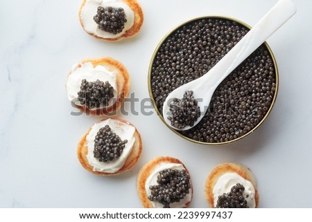 Black caviar in the can with a pearl spoon and appetizers mini pancakes with cream, close up top view, Horizontal on a white marble background with a copy space