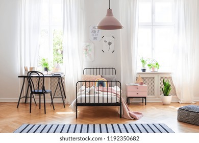 Black Chair At Desk Next To Bed In Girl's Room Interior With Pink Lamp Above Carpet. Real Photo