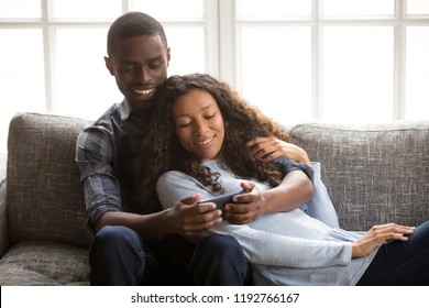 Black Cheerful Married Couple In Love Sitting Together On Couch In Living Room Or Hotel Using Mobile Phone. Happy Husband And Joyful Wife Spend Free Time On Weekend Watching Funny Recreational Video