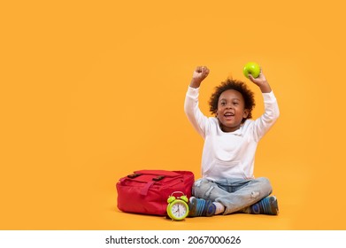 Black Child Boy 3 Years, Student Kid Holding Green Apple In His Hand And Yay! Happy While Sitting With School Bag And Alarm Clock. Isolated Portrait On Yellow Background With Copy Space.