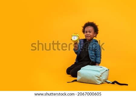 Black child boy 3 years, Student kid holding an alarm clock in his hand while sitting with school bag. Isolated portrait on yellow background with copy space. International students