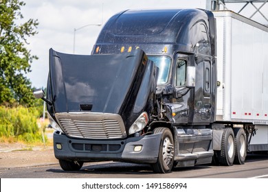 Black Classic American Bonnet Big Rig Semi Truck With Open Hood Standing On The Roadside For Waiting For A Mobile Towing For Repair Engine And Schedule Maintenance Service