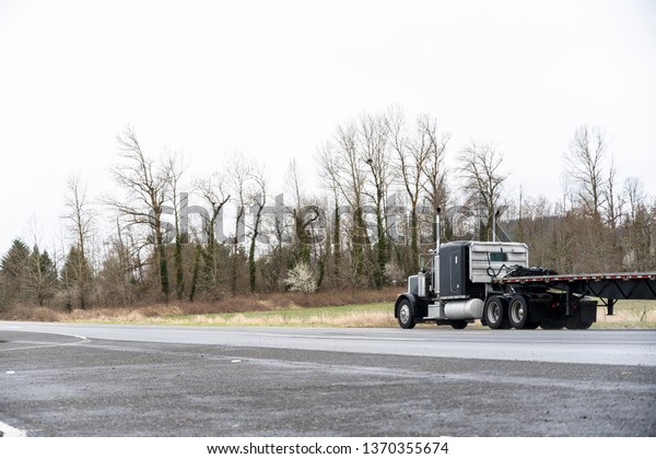 Black classic American idol big rig semi truck with\
chrome with vertical tailpipes transporting empty flat bed semi\
trailer on the road with bare trees going to warehouse for loading\
commercial cargo