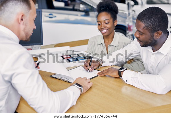 black clients sign a contract while\
sitting at table with salesman, professional worker of dealership\
explain terms of the contract and show where to\
sign