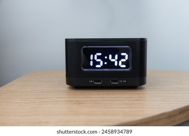 A black clock with the time 15:42 on it sits on a wooden table. The clock is small and has a black face with white numbers. The time is displayed in a clear and easy-to-read manner