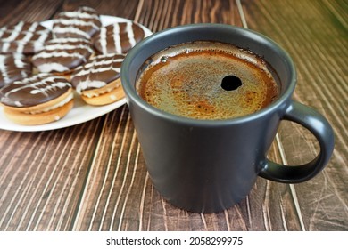 A Black Coffee Mug Sits On A Wooden Table Next To A White Plate Of Chocolate Chip Cookies. Side View