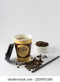 Black Coffee In A Tall Cardboard Glass On A White Table. Coffee Beans In A White Vase In The Background. A Black Straw On The Table. White Background, Text Space