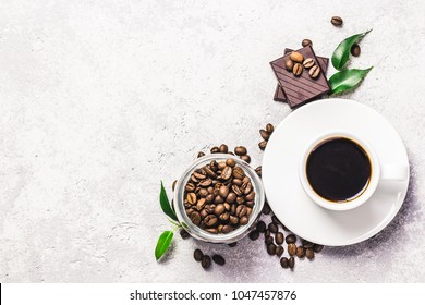 Black coffee in white cup, chocolate and coffee beans in glass jar on concrete background. Top view, space for text.