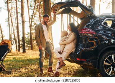 With black colored automobile. Happy family of father, mother and little daughter is in the forest.