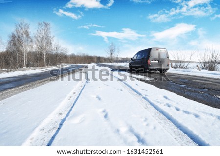 Black compact van on the winter countryside road with snow against blue sky with clouds