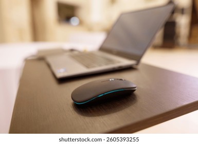 A black computer mouse sits on a wooden desk next to a laptop. The mouse is black and has a green stripe. The laptop is open and turned on